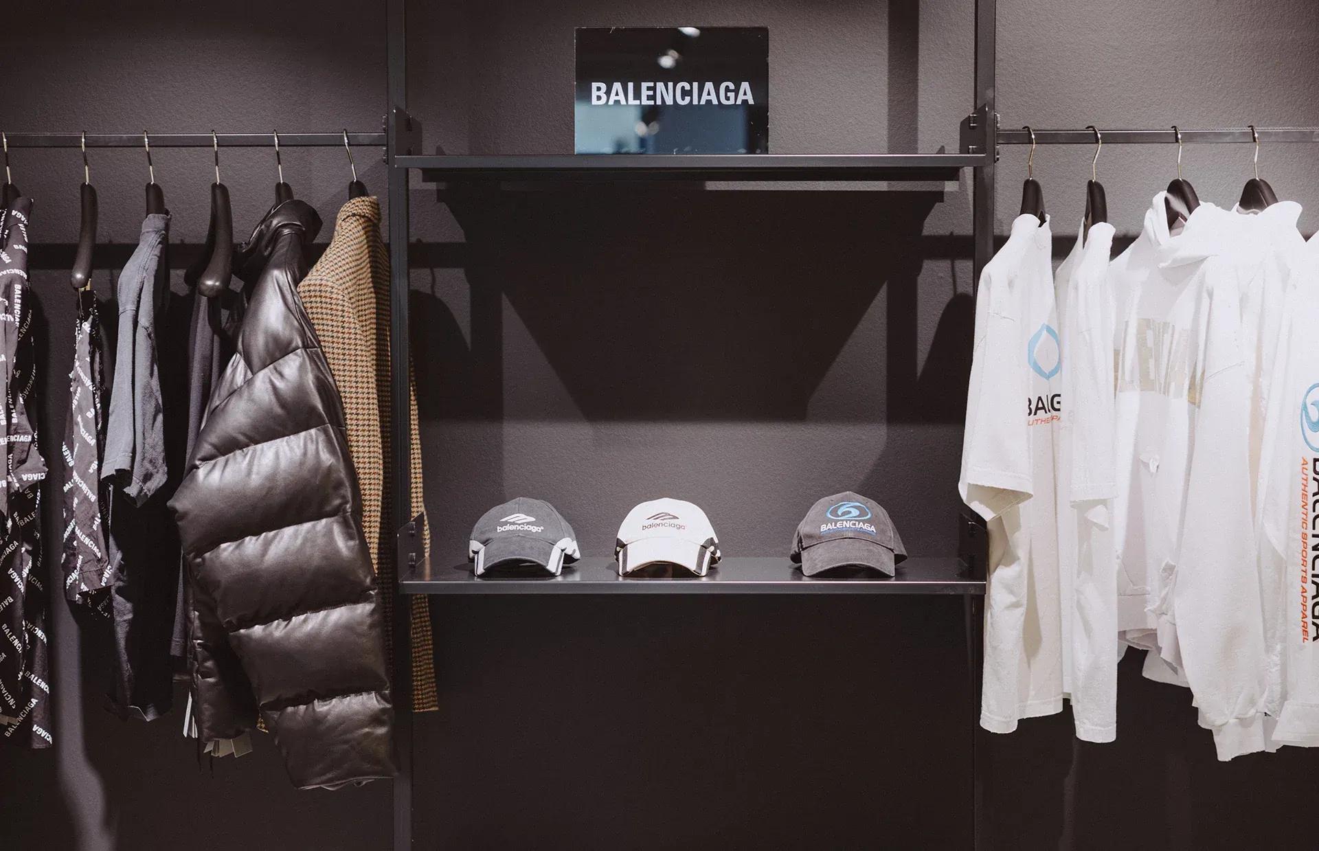 Clothing rack with Balenciaga branded items including a black puffer jacket and white t-shirts.