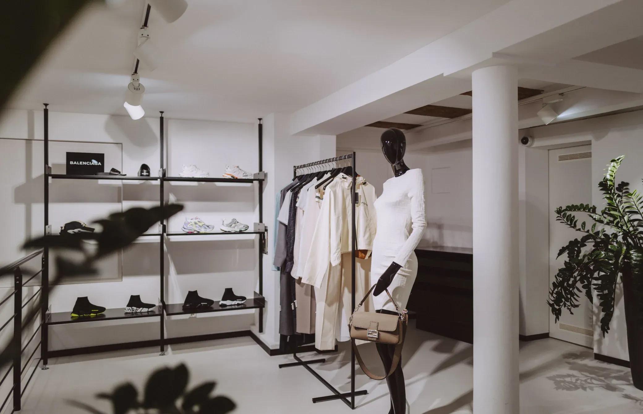 Bright clothing store interior with a mannequin dressed in white, near black racks and a plant.