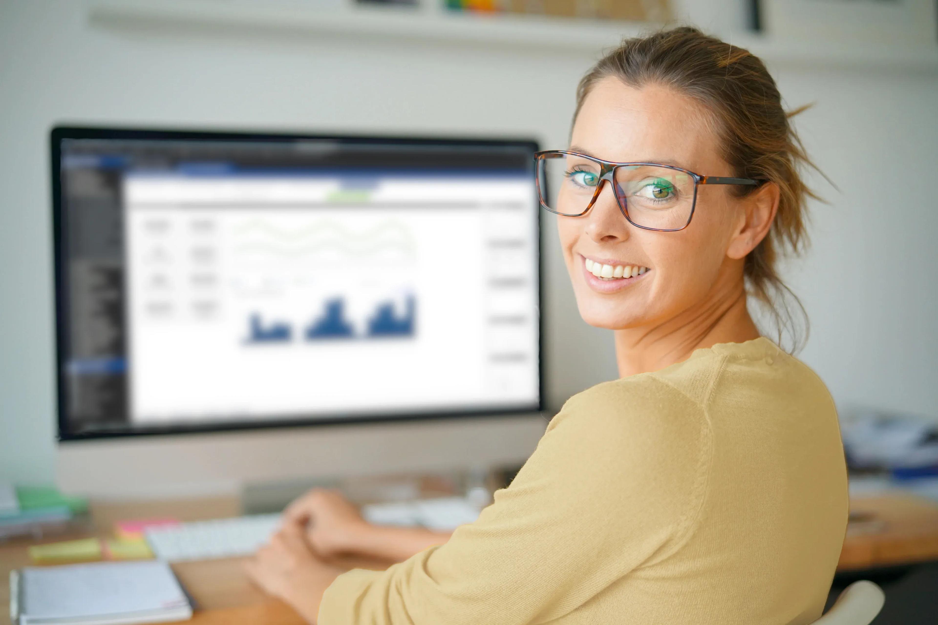 Smiling woman in glasses looks over shoulder at camera while working on computer.