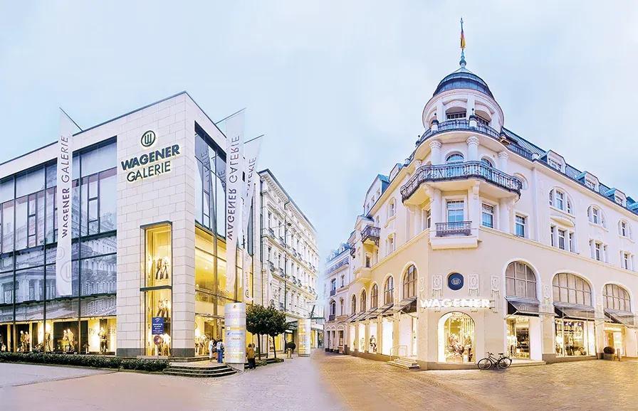 Two large, elegant city buildings, one labeled "WAGNER GALERIE" under a cloudy sky.