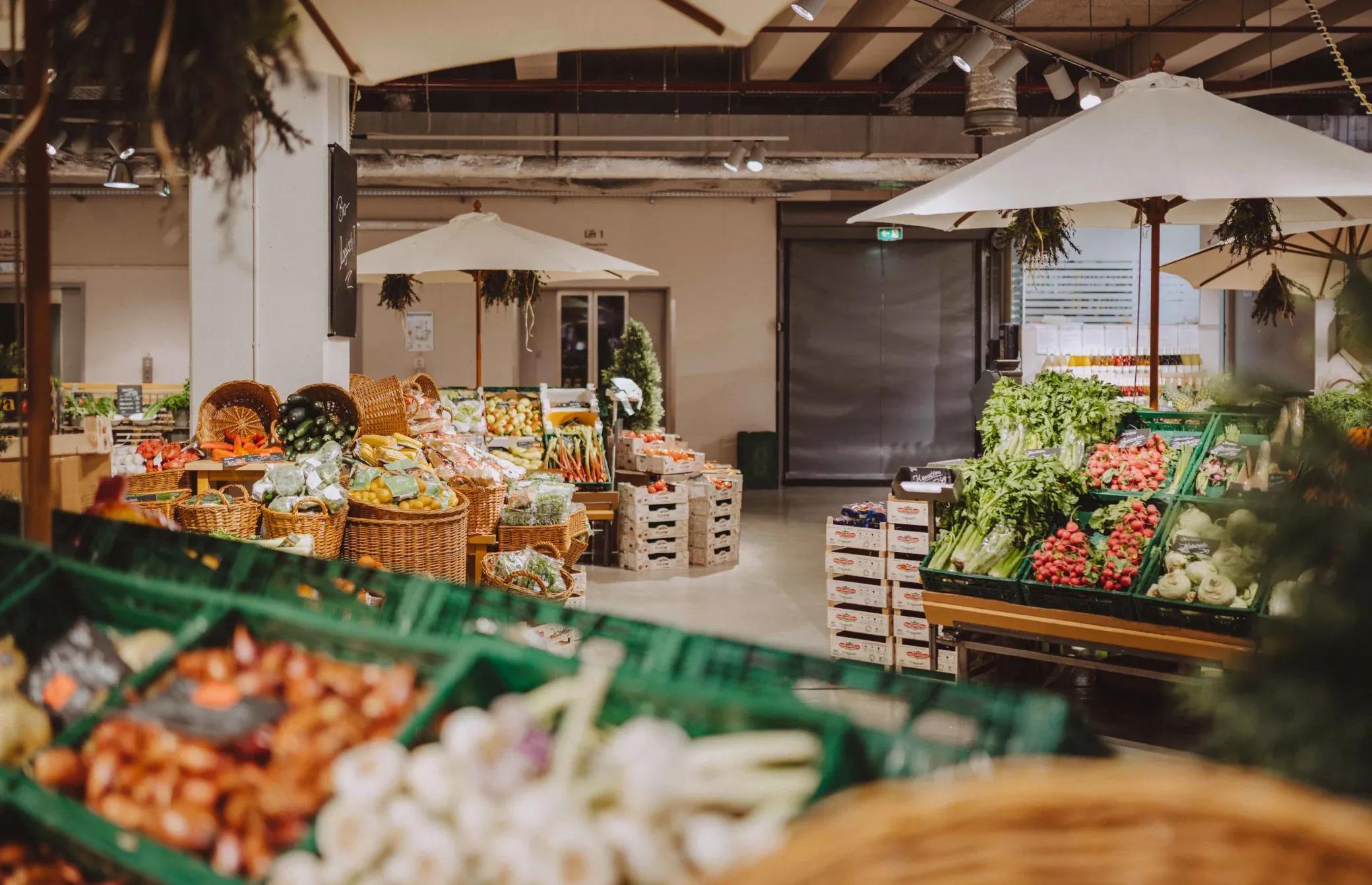 Indoor market displaying fresh fruits and vegetables in wooden crates under market umbrellas.