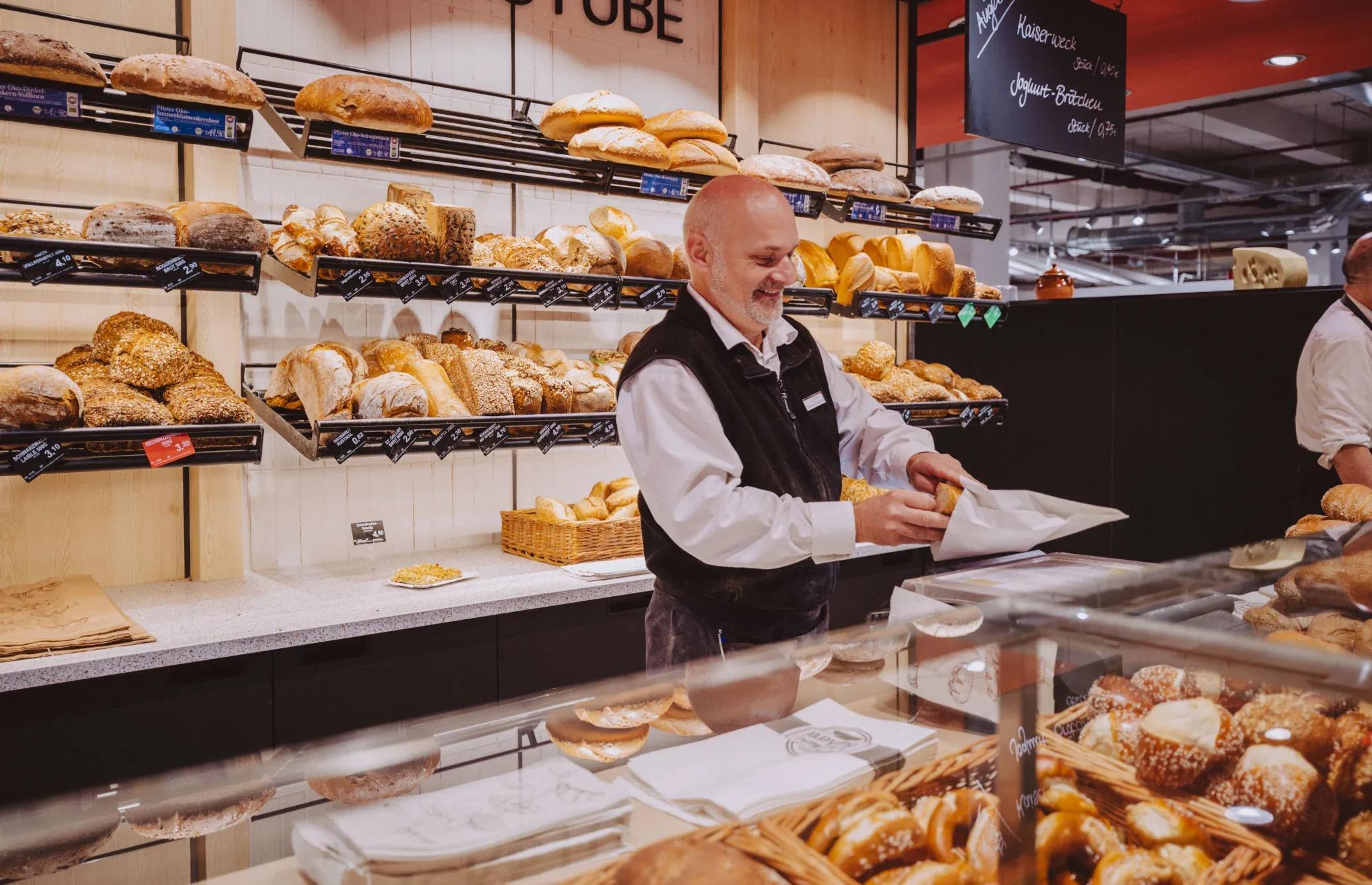 Smiling baker puts freshly baked bread into a paper bag at a well-stocked bakery.