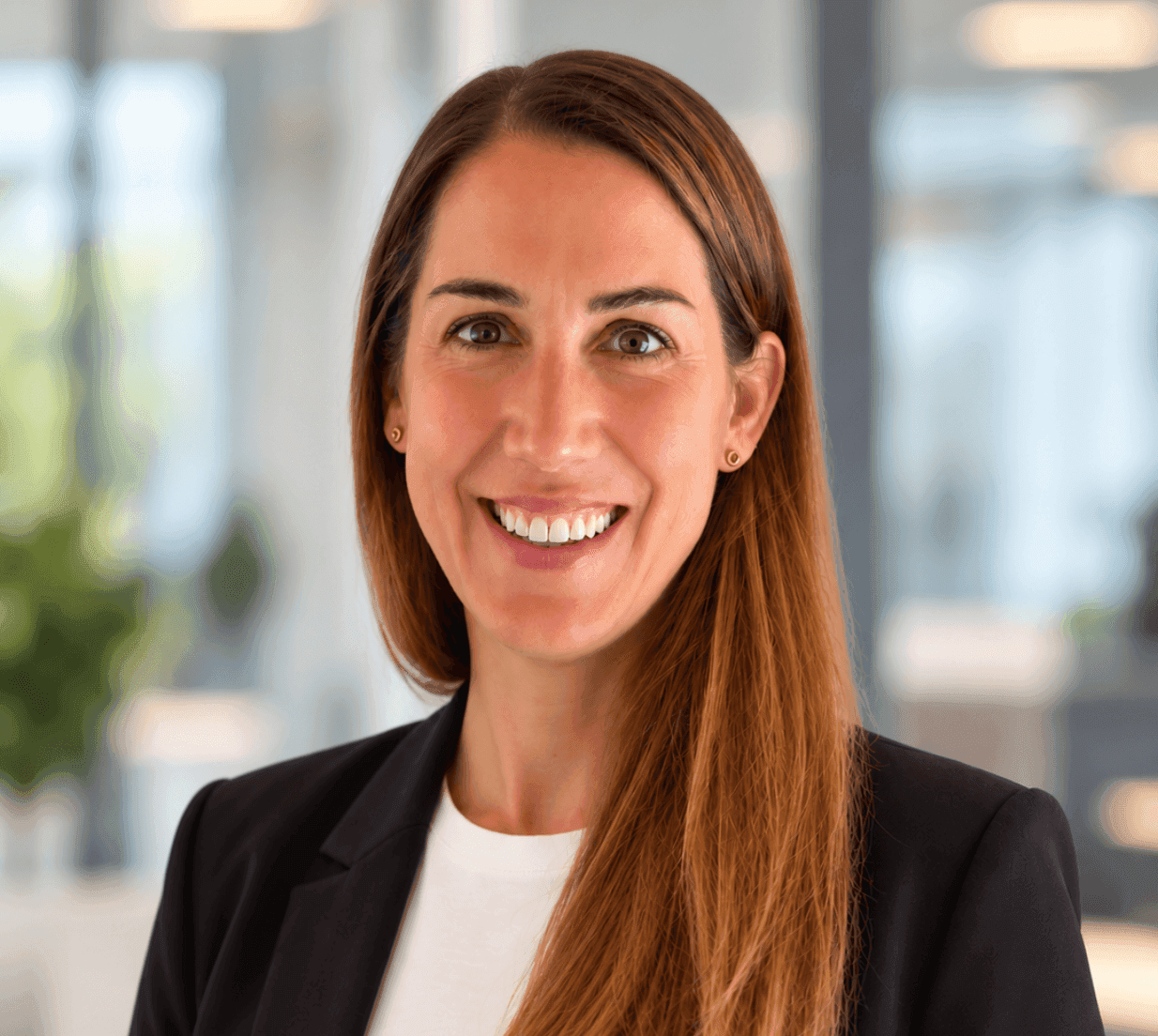 Smiling woman with long brown hair wearing a black blazer in a modern office environment.